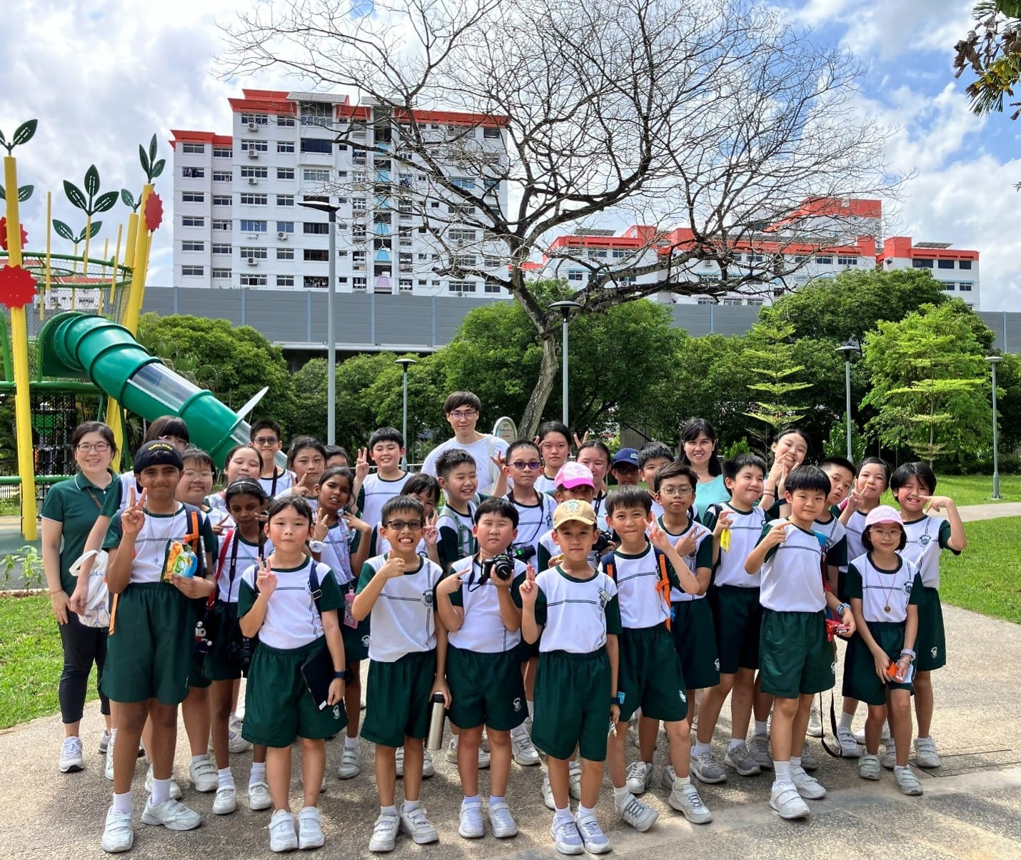 Media Club students and teachers posing cheerfully for a group photo at a neighbourhood park during an outdoor photography excursion, with a playground and HDB residential blocks visible in the background.